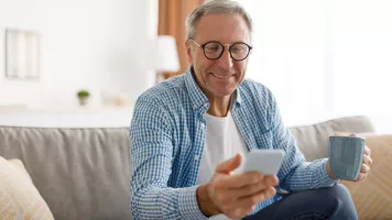 Hombre blanco de pelo gris con gafas en el sofá sosteniendo una taza de café y mirando un teléfono