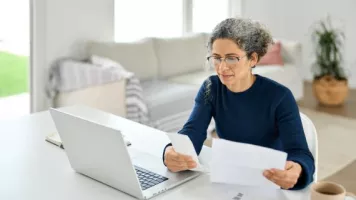 Una señora mayor blanca con una camisa oscura mirando papeles y una computadora