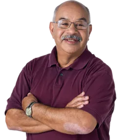 Hombre hispano mayor con anteojos y bigote gris. Tiene los brazos cruzados de forma relajada y lleva un polo granate de manga corta y un reloj. 